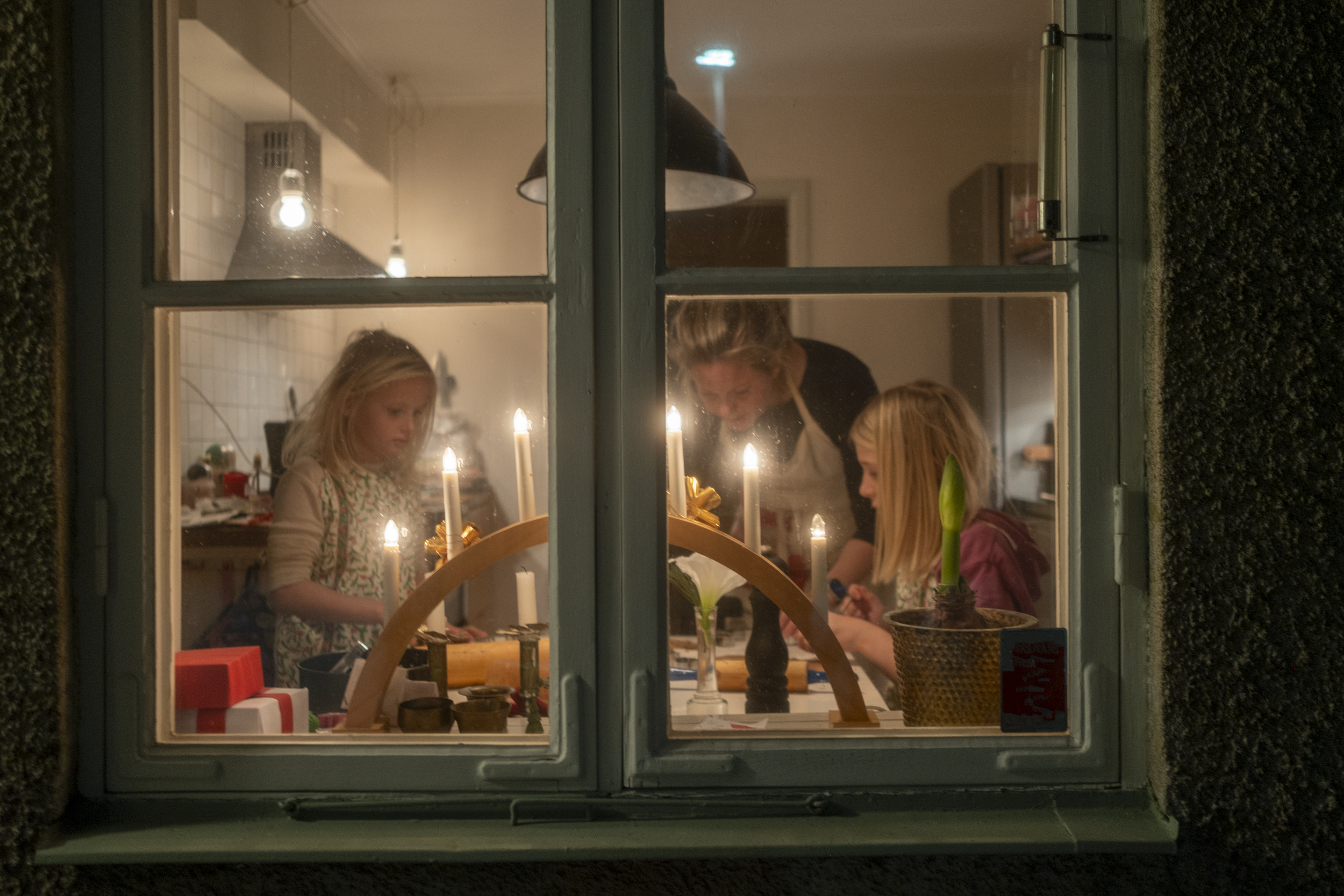 Gjennom et vindu ser vi en kvinne og to barn inne på et kjøkken. De holder på med juleforberedelser.